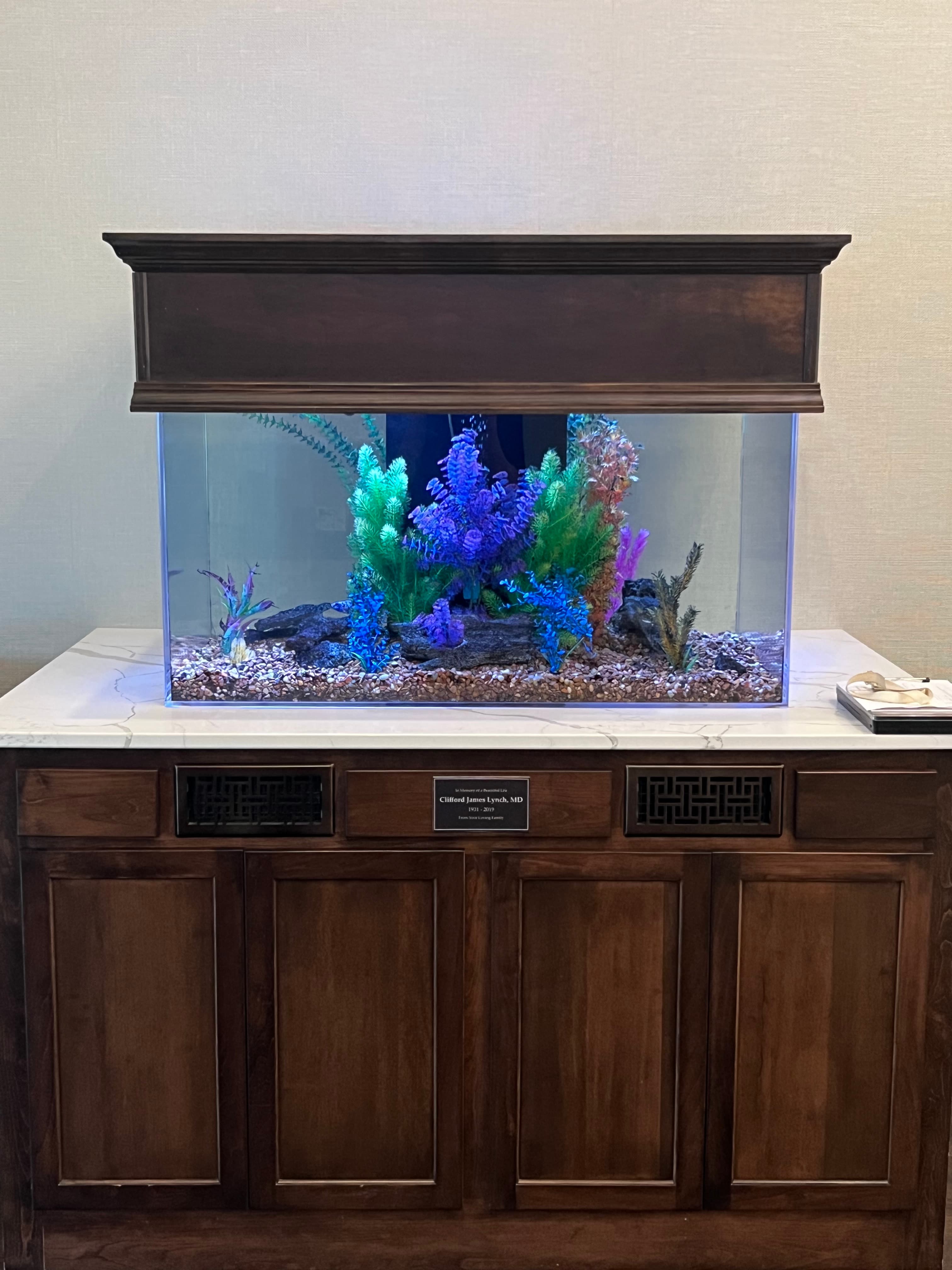 Vibrant aquarium with colorful plants built into a dark wood memorial cabinet with a plaque.